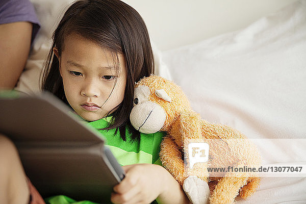 Mädchen mit Plüschtier benutzt Tablet-Computer  während sie neben der Mutter zu Hause im Bett liegt