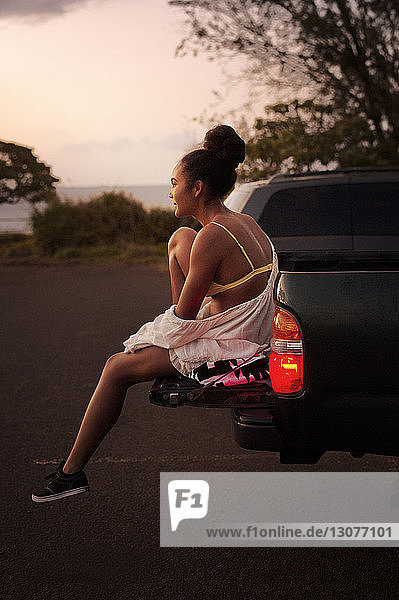 Side view of teenager sitting on truck bed liner parked on road during sunset