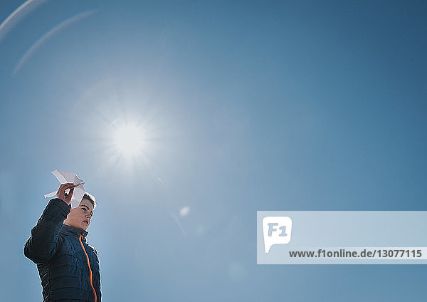 Niedrigwinkelansicht eines Jungen  der ein Papierflugzeug hält  während er an einem sonnigen Tag vor einem klaren blauen Himmel steht