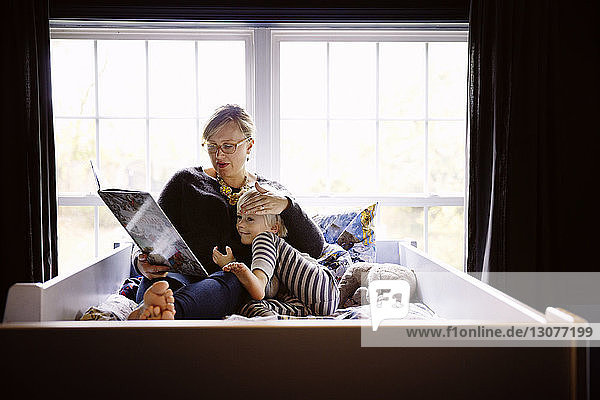 Mother reading book to son while sitting on bed against window at home