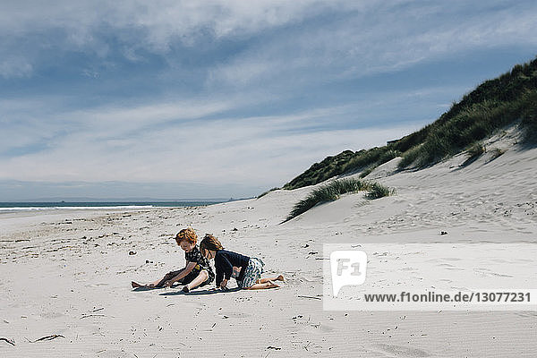 Geschwister in voller Länge beim Spielen mit Sand am Strand vor bewölktem Himmel am sonnigen Tag