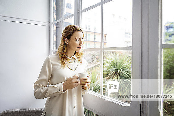 Geschäftsfrau schaut durch ein Fenster  während sie eine Kaffeetasse im Büro hält
