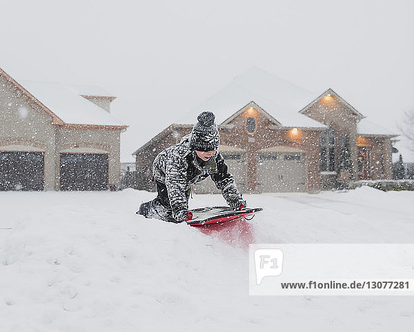 Boy tobogganing on snow against houses during snowfall