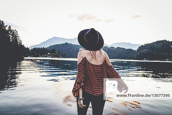 Rear view of woman if fedora hat looking at view while standing by lake during dusk