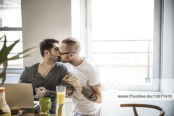 Homosexual couple kissing while having breakfast at table against window