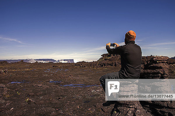 Rear view of male hiker photographing while sitting on rock against clear blue sky