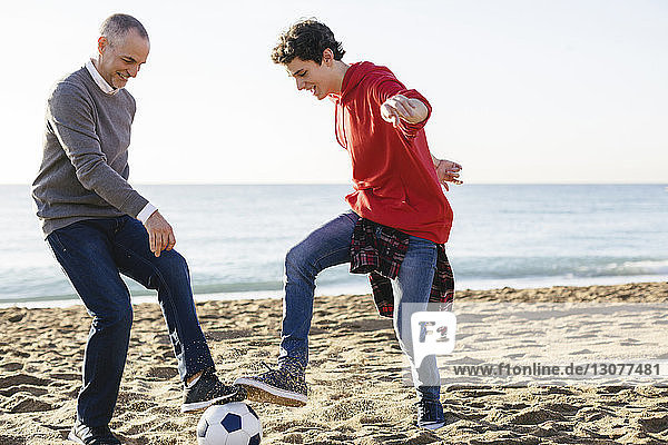 Playful father and son playing soccer at beach against clear sky
