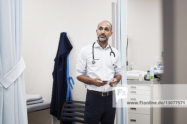 Doctor with smart phone looking away while standing in room at hospital