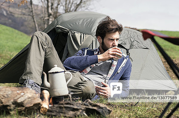 Nachdenklicher Mann trinkt Tee  während er sich vor dem Zelt auf dem Hügel entspannt