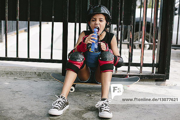 Verspieltes Mädchen trinkt Wasser  während es auf einem Skateboard gegen ein Geländer sitzt