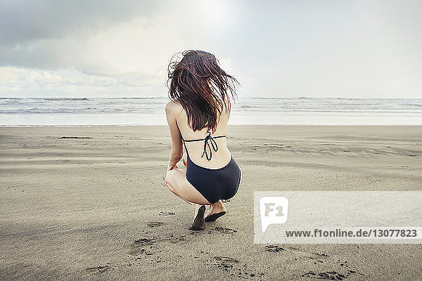 Rear view of woman crouching at Bethells Beach against sea and sky