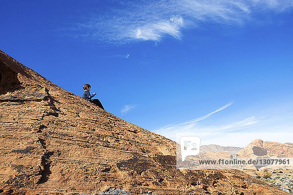 Low angle view of boy sitting on rock at Valley of Fire State Park against blue sky