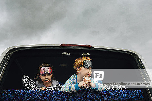 Geschwister mit Schlafmasken im Fahrzeug liegend  während sie gegen bewölkten Himmel wegschauen