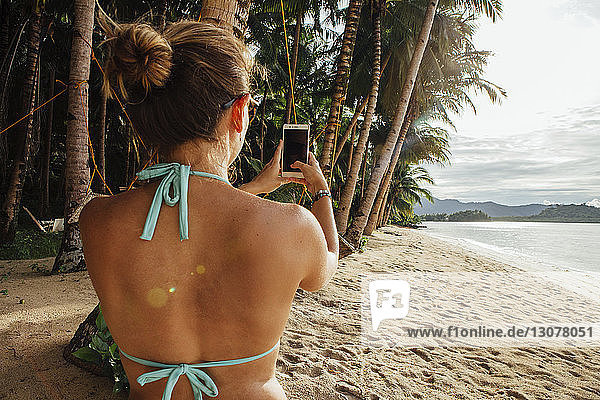 Rückansicht einer jungen Frau  die ein Smartphone am Strand benutzt