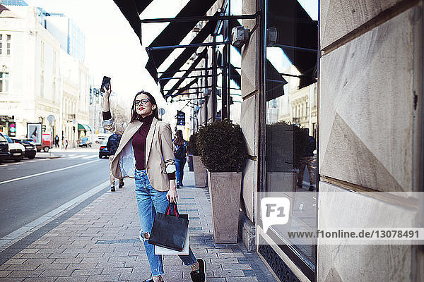 Frau mit Einkaufstaschen und Mobiltelefon auf dem Bürgersteig in der Stadt stehend