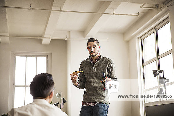 Businessman having pizza and beer while talking to colleague at creative office