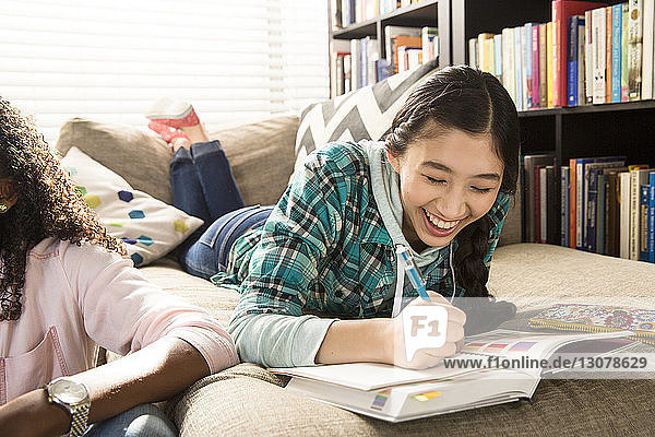 Fröhliches Teenagermädchen macht Hausaufgaben  während es zu Hause bei einem Freund auf der Couch liegt