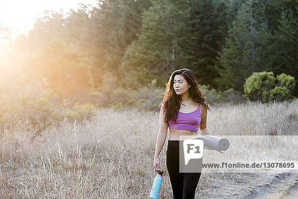 Woman holding exercise mat and water bottle while walking at field