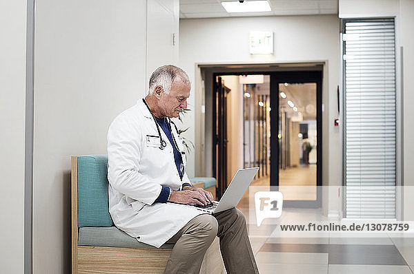 Senior doctor using laptop computer while sitting on sofa in corridor