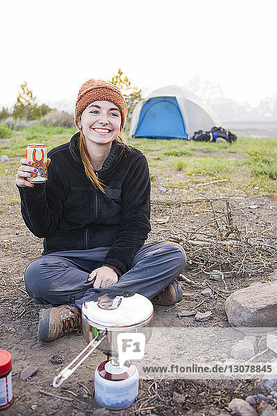 Lächelnde Frau hält Getränk in der Hand  während sie auf dem Campingplatz sitzt