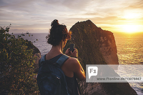 Rear view of man with backpack photographing sea while standing on mountain during sunset