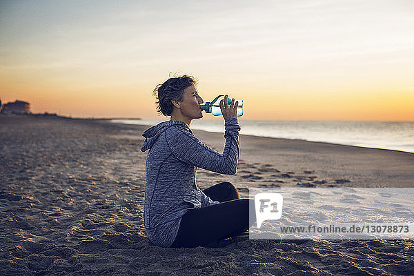 Frau trinkt Wasser  während sie am Strand gegen den Himmel sitzt