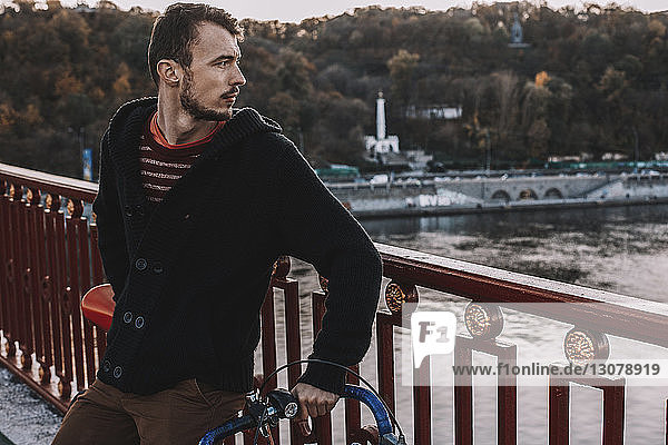 Thoughtful man looking away while standing with bicycle on bridge over river against trees during sunset