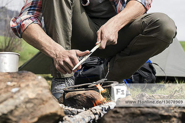Niedriger Abschnitt eines Mannes  der einen Stock über einem Lagerfeuer auf einem Campingplatz schneidet