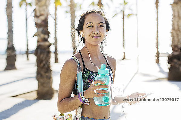 Porträt einer glücklichen sportlichen Frau  die im Sommer auf einem Fußweg eine Wasserflasche hält