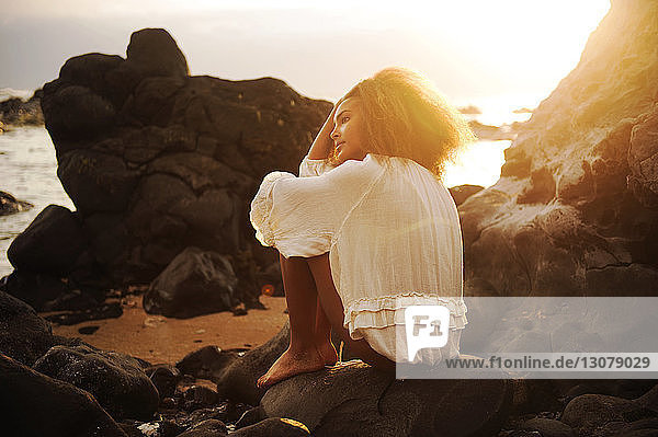 Nachdenkliches Teenager-Mädchen sitzt bei Sonnenschein auf einem Felsen am Meeresufer