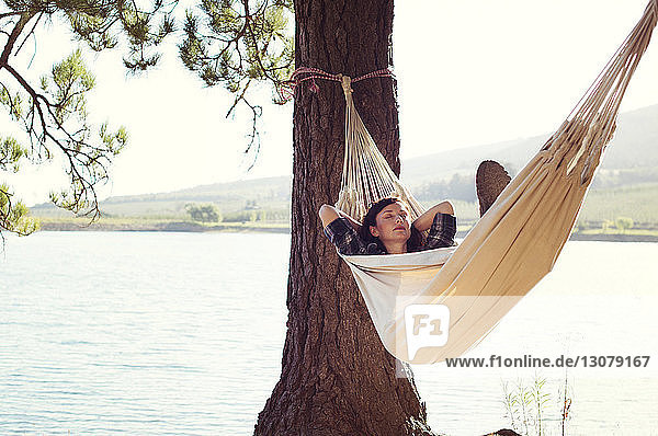 Woman sleeping on hammock by tree at lakeshore