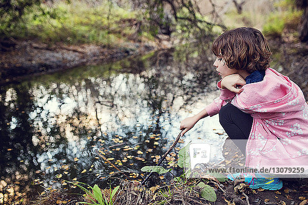 Seitenansicht eines Mädchens  das einen Stock hält  während es am Bach im Wald sitzt