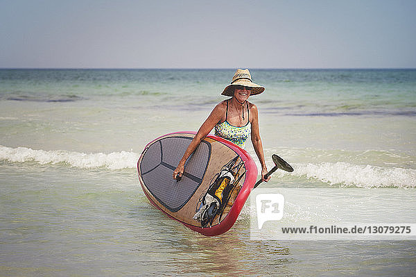 Woman carrying paddleboard while walking in sea against clear sky