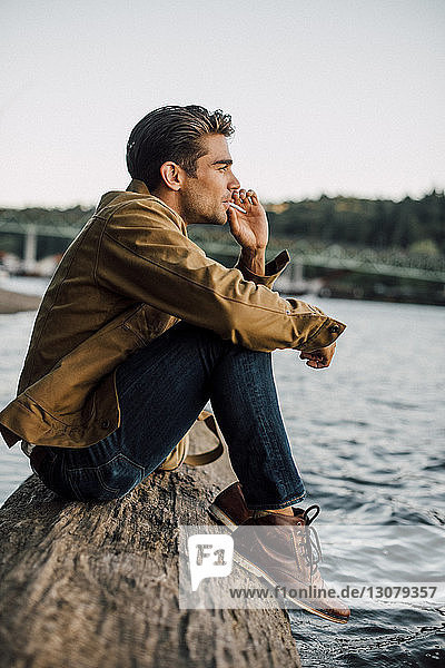 Man smoking while sitting on log by river against clear sky