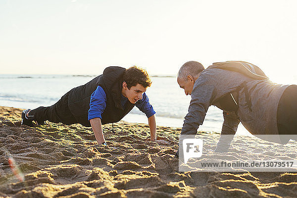 Determined father and son doing push-ups together against clear sky at beach during sunset
