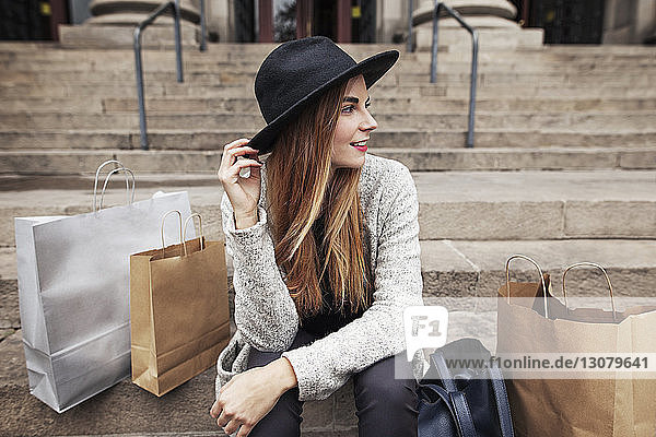 Glückliche Frau mit Hut  die auf der Treppe neben Einkaufstaschen sitzt