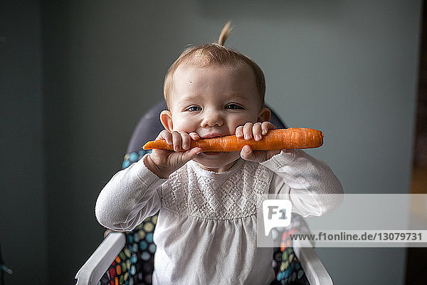 Porträt eines süßen Mädchens beißt in Karotten  während es zu Hause auf einem Hochstuhl sitzt