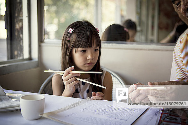 Grandfather teaching granddaughter to hold chopsticks while sitting in restaurant