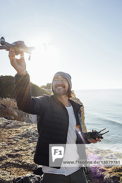 Smiling man flying quadcopter while standing at beach against clear sky during sunny day