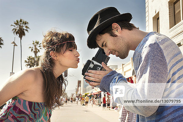 Seitenansicht einer Frau  die die Lippen kräuselt  während ein Mann durch eine alte Kamera auf dem Bürgersteig fotografiert
