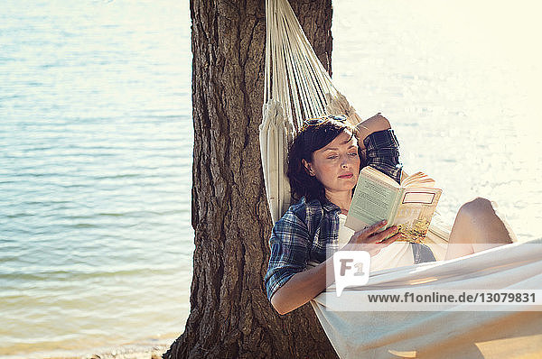 Woman reading book while relaxing on hammock by tree at lakeshore