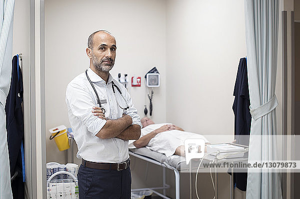 Porträt eines selbstbewussten Arztes  der im medizinischen Untersuchungsraum mit dem Patienten im Hintergrund steht