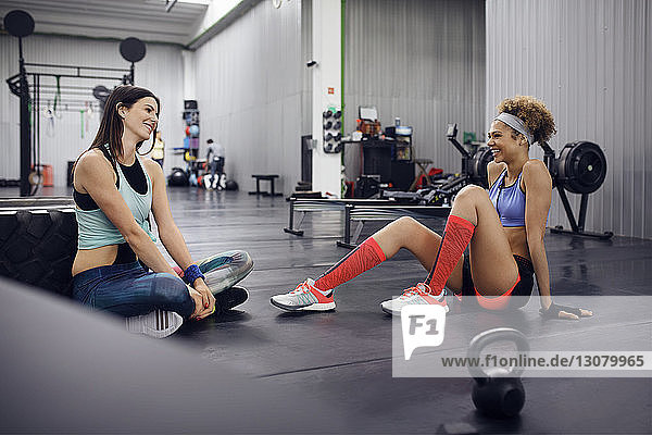 Glückliche Sportlerinnen unterhalten sich  während sie im Fitnessstudio auf dem Boden sitzen