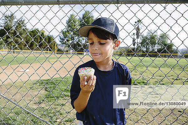 Boy eating cupcake while standing by chainlink fence at playing field