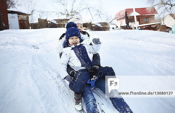 Cheerful mother and daughter tobogganing on snowy field