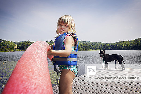 Porträt eines Mädchens  das ein Nudelschwimmen hält  während es mit einem Hund auf dem Pier steht