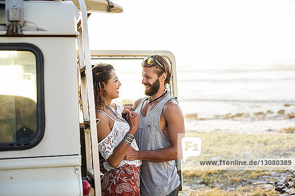 Cheerful couple standing by off-road vehicle at beach against clear sky