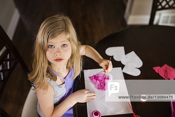 Hochwinkelporträt eines Mädchens  das zu Hause Herzformen aus Papieren auf dem Tisch herstellt