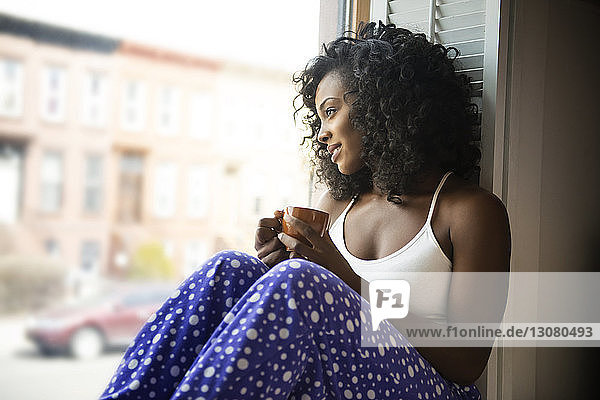 Lächelnde Frau schaut durchs Fenster  während sie zu Hause Kaffee trinkt