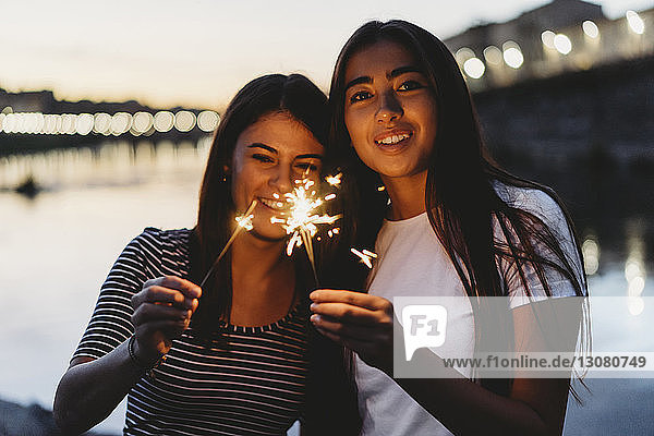 Happy friends holding burning sparklers while sitting by Arno river against sky during sunset